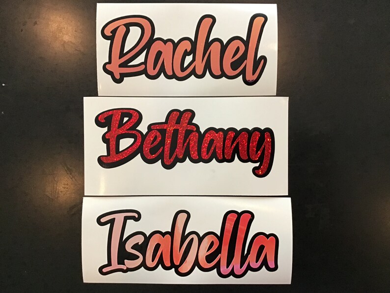 May include: Three white vinyl stickers with names written in cursive. The names are Rachel, Bethany, and Isabella. The names are written in different shades of red and pink with black outlines.