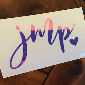 May include: A white sticker with the word "jump" in a stylized script, with the letters in purple and pink. A small purple heart is next to the last letter. The sticker is on a wooden surface.