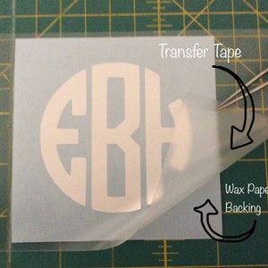 May include: A white vinyl monogram sticker with the letters "EBH" in a circle, on a clear transfer tape sheet. The transfer tape is being peeled off of the wax paper backing.