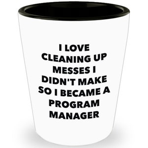 May include: White ceramic shot glass with a black rim. The glass has black text that reads "I love cleaning up messes I didn't make so I became a program manager."