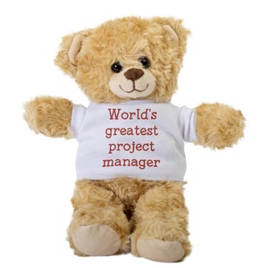 May include: A plush teddy bear with light brown fur and a white t-shirt. The t-shirt has the text "World's greatest project manager" in a dark red color. The bear has a friendly expression.