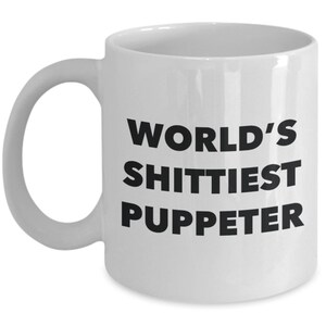 May include: White ceramic coffee mug with black text that reads "WORLD'S SHITTIEST PUPPETER".