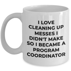 May include: White ceramic coffee mug with black text that reads "I love cleaning up messes I didn't make so I became a program coordinator."