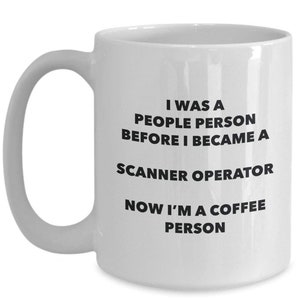Könnte beinhalten: Weiße Keramik-Kaffeetasse mit gebogenem Henkel. Die Tasse hat einen schwarzen Text, der lautet: "I WAS A PEOPLE PERSON BEFORE I BECAME A SCANNER OPERATOR NOW I'M A COFFEE PERSON."