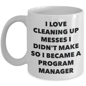 May include: White ceramic coffee mug with black text that reads "I love cleaning up messes I didn't make so I became a program manager".