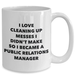 Könnte beinhalten: Eine weiße Keramik-Kaffeetasse mit schwarzem Text, der lautet: "Ich liebe es, Unordnung zu beseitigen, die ich nicht verursacht habe, deshalb bin ich PR-Manager geworden."