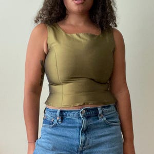 Pode incluir: Top sem mangas verde-oliva com decote quadrado e cintura justa. A parte superior é feita de um tecido brilhante. A modelo está usando jeans azuis. A parte superior é uma adição elegante a qualquer guarda-roupa.