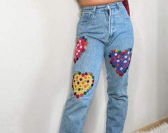 May include: Light blue denim jeans with three embroidered heart patches. Each heart features a different color scheme of flowers, including pink, yellow, and purple. The jeans have a classic five-pocket design and a button and zipper closure.