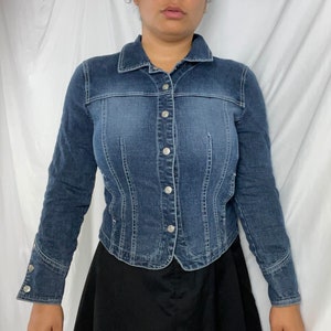May include: A fitted, dark-wash denim jacket with a button-up front and long sleeves. The jacket has a classic collar and silver-colored buttons. The design includes vertical seams for a tailored fit. The jacket is worn over a black skirt.