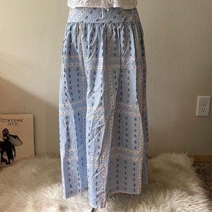 Pu&ograve; includere: Un completo a due pezzi su un manichino. Il top &egrave; una camisole bianca traforata con un davanti allacciato. La gonna &egrave; una gonna lunga azzurra con un motivo ripetuto di piccoli disegni floreali neri e bianchi. &Egrave; visibile anche una collana con pendente argentato.