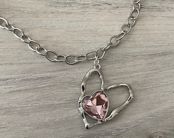 May include: A silver chain necklace with a heart-shaped pendant. The pendant has a pink crystal in the center.