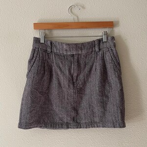 May include: A gray herringbone mini skirt hanging on a wooden hanger. The skirt has a classic design with a zipper and pockets. The fabric has a textured appearance.