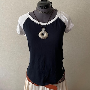 Puede incluir: Una camiseta de béisbol azul marino y blanca con un collar colgante plateado grande. El collar tiene un diseño en espiral. La camisa se muestra en un maniquí negro. Se ve un cinturón marrón.