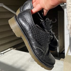 Puede incluir: Zapatos Oxford de cuero negro con textura de piel de cocodrilo sint&eacute;tica. Los zapatos tienen un dise&ntilde;o con cordones y una suela gruesa de color marr&oacute;n claro. Los zapatos est&aacute;n sostenidos por una mano.