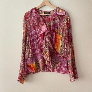 Könnte beinhalten: Ein langärmeliges Hemd mit Knopfleiste und einem lebendigen Patchwork-Muster in Rosa-, Orange-, Gelb- und Violetttönen. Das Hemd verfügt über eine gerüschte Knopfleiste, einen klassischen Kragen und ein Spitzenbesatz-Detail in der Mitte.