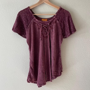 Pode incluir: Uma blusa bordô de manga curta com detalhes de renda nas mangas e decote. A blusa tem uma frente com cadarço e está pendurada em um cabide de madeira. O tecido tem uma aparência texturizada.