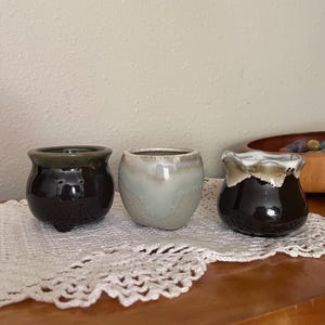 May include: Three small ceramic pots in varying colors: dark brown with a green rim, light blue-gray, and dark brown with a white and brown rim. The pots are round with a small base and sit on a white lace doily.