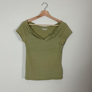 May include: Olive green short-sleeve shirt with a V-neck and a gathered neckline detail. The shirt is hanging on a wooden hanger against a plain white background. The shirt is made of a soft, lightweight material.