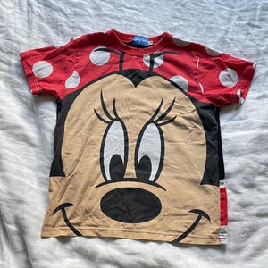 May include: A red t-shirt with white polka dots on the sleeves and a large Minnie Mouse graphic on the front. The graphic is in black, tan, and white. The t-shirt has a round neck and short sleeves.