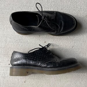 Puede incluir: Par de zapatos de cuero negro con textura de piel de cocodrilo. Los zapatos tienen un dise&ntilde;o con cordones y una suela gruesa de color marr&oacute;n claro. Los zapatos se muestran sobre una superficie gris claro.