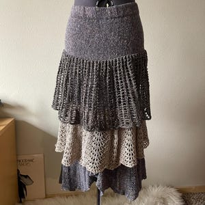 May include: A tiered skirt in shades of gray and beige, displayed on a mannequin. The top section is a solid gray knit, followed by a dark gray openwork tier, a beige openwork tier, and a final dark gray tier. The skirt has a bohemian style.