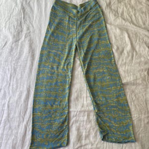 May include: Full-length trousers featuring a high-waisted design and a wide-leg silhouette. The fabric displays a repeating abstract pattern in shades of light blue and olive green, creating a textured visual effect across the entire garment.