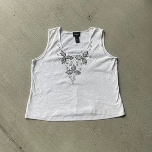 May include: A white sleeveless tank top with a beaded floral design on the front. The tank top has a scoop neck and is made of a soft, lightweight fabric.