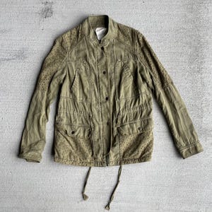 Pode incluir: Casaco verde-oliva com gola alta, fecho de bot&otilde;es e v&aacute;rios bolsos. O casaco apresenta detalhes perfurados nos ombros e na parte inferior da frente. Cord&otilde;es s&atilde;o vis&iacute;veis na cintura e na bainha. Uma pe&ccedil;a de roupa exterior casual.