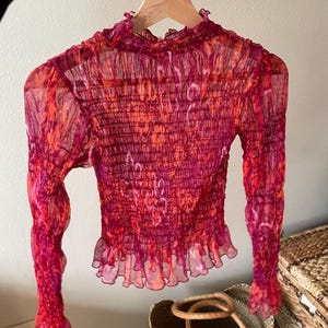 May include: A sheer, long-sleeved top in shades of red and orange, hanging on a wooden hanger. The top features a high neckline, a ruched bodice, and a ruffled hemline. The fabric has a subtle, patterned design.