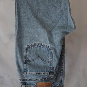 Puede incluir: Un par de pantalones vaqueros de mezclilla azul claro colgados de una percha de madera. Los pantalones tienen un bolsillo trasero con un detalle de costura roja y un parche de cuero marr&oacute;n con el texto "Levi's" y "Original  Rivet &  Clothing Co.  San Francisco,  Ca. 90210".
