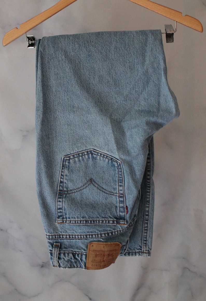 Puede incluir: Un par de pantalones vaqueros de mezclilla azul claro colgados de una percha de madera. Los pantalones tienen un bolsillo trasero con un detalle de costura roja y un parche de cuero marr&oacute;n con el texto "Levi's" y "Original  Rivet &  Clothing Co.  San Francisco,  Ca. 90210".