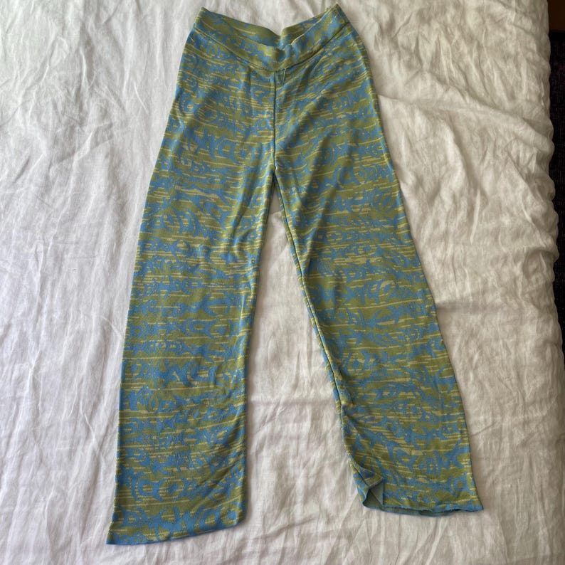 May include: Full-length trousers featuring a high-waisted design and a wide-leg silhouette. The fabric displays a repeating abstract pattern in shades of light blue and olive green, creating a textured visual effect across the entire garment.