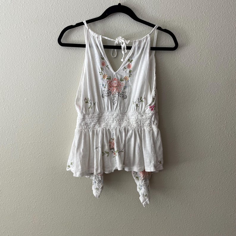May include: White tank top with floral embroidery in pink, green, and peach. The top has a V-neck, a gathered waist, and tie closures at the bottom. The top is hanging on a black hanger.
