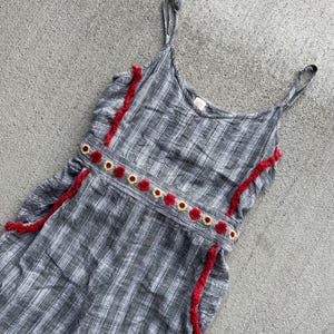 K&ouml;nnte beinhalten: Ein gestreiftes Sommerkleid in Grau und Wei&szlig; mit Spaghettitr&auml;gern. Das Kleid hat rote Fransenakzente und eine Reihe von rot-gelben, gestickten Blumen auf dem Oberteil. Der Stoff hat ein strukturiertes Aussehen, und das Kleid liegt flach auf einer grauen Oberfl&auml;che.