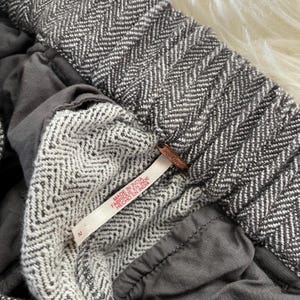 May include: Close-up of gray and white herringbone patterned fabric, possibly pants or a skirt. The garment has a tag that reads "MADE IN INDIA". The fabric is a mix of gray and white, with a textured appearance. The garment is size 2.