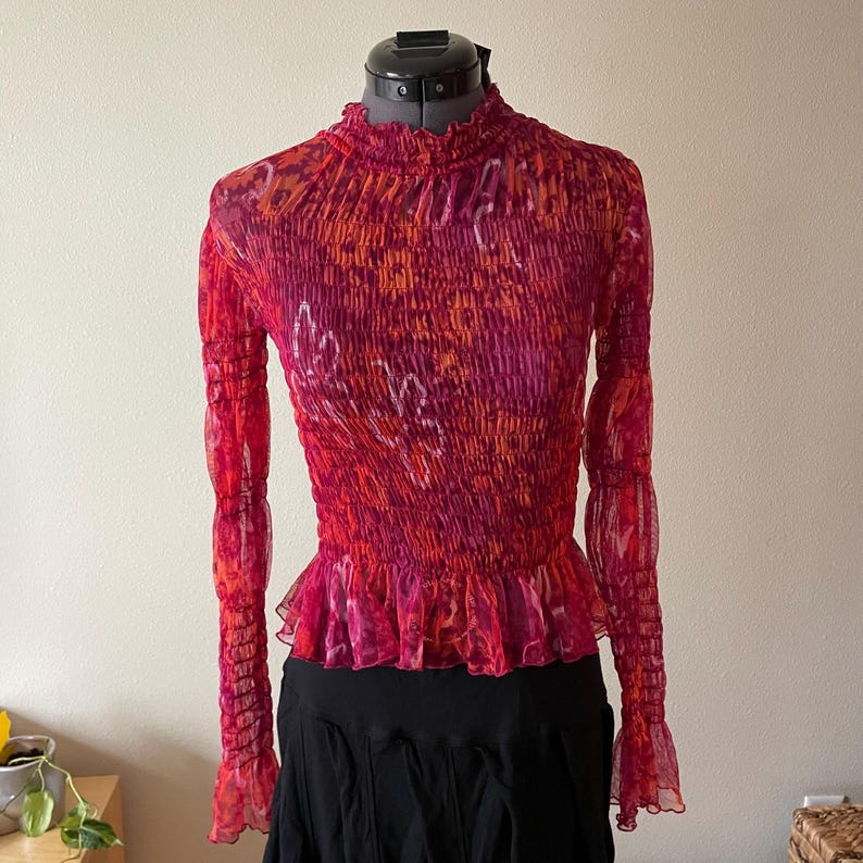 May include: A long-sleeved, red, orange, and pink patterned top with a high neckline and ruffled hem. The top has a fitted bodice and sheer sleeves with ruffled cuffs. The garment is displayed on a mannequin.