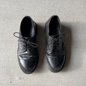 Puede incluir: Un par de zapatos de vestir de cuero negro con un patr&oacute;n texturizado similar al de un cocodrilo. Los zapatos tienen un dise&ntilde;o cl&aacute;sico con cordones y una puntera redondeada. Las suelas son oscuras y los zapatos parecen desgastados.