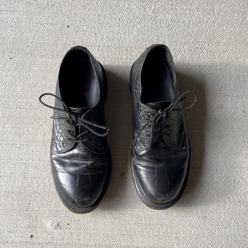 Puede incluir: Un par de zapatos de vestir de cuero negro con un patr&oacute;n texturizado similar al de un cocodrilo. Los zapatos tienen un dise&ntilde;o cl&aacute;sico con cordones y una puntera redondeada. Las suelas son oscuras y los zapatos parecen desgastados.