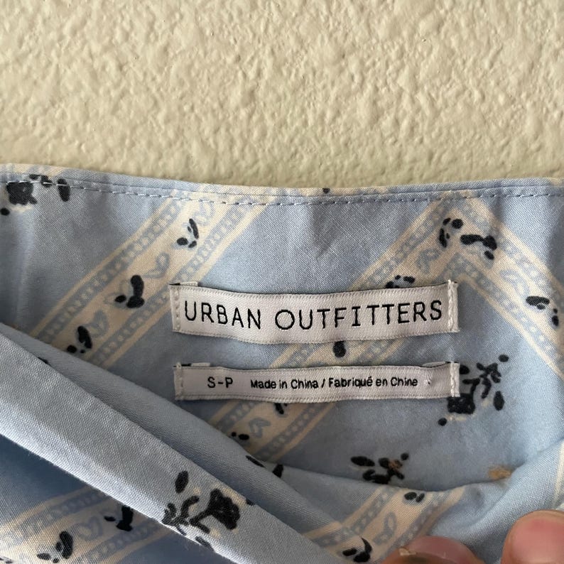 Pu&ograve; includere: Primo piano di un tessuto azzurro con un motivo paisley e l'etichetta del marchio "URBAN OUTFITTERS". Il tessuto presenta un motivo ripetuto di strisce bianche e accenti floreali neri. Taglia S-P.