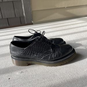 Puede incluir: Un par de zapatos de cuero negro con un patr&oacute;n texturizado similar al de un cocodrilo. Los zapatos tienen un dise&ntilde;o con cordones y una suela gruesa y oscura. Los zapatos est&aacute;n sobre una superficie de hormig&oacute;n.