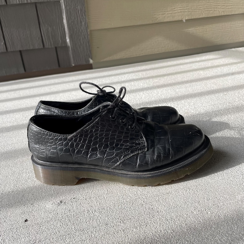 Puede incluir: Un par de zapatos de cuero negro con un patr&oacute;n texturizado similar al de un cocodrilo. Los zapatos tienen un dise&ntilde;o con cordones y una suela gruesa y oscura. Los zapatos est&aacute;n sobre una superficie de hormig&oacute;n.
