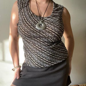 May include: Sleeveless top with a repeating wave pattern in shades of brown and cream. The top is paired with a dark gray skirt. A silver pendant hangs from a black cord necklace. A gold bracelet and a gold bangle are visible.