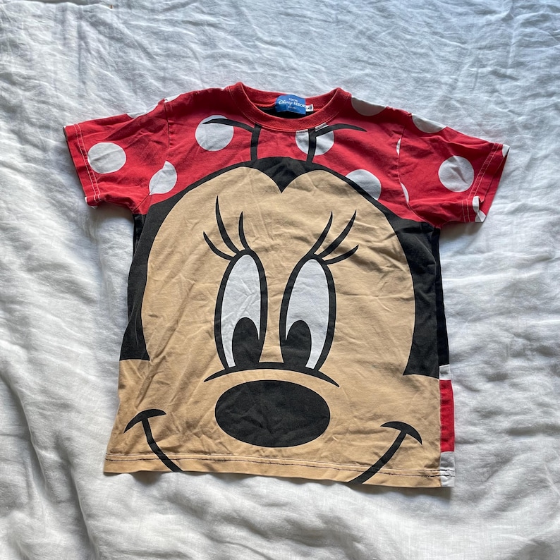 May include: A red t-shirt with white polka dots on the sleeves and a large Minnie Mouse graphic on the front. The graphic is in black, tan, and white. The t-shirt has a round neck and short sleeves.