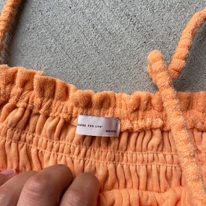 Pode incluir: Um top halter cor p&ecirc;ssego, canelado, com decote franzido e uma pequena etiqueta que diz "GOODS FOR LIFE" e "MADE IN CHINA".