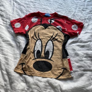 May include: A red t-shirt with white polka dots and a large Minnie Mouse graphic. The shirt features a tan face, black eyes, and a black nose. The sleeves are red with white polka dots. The t-shirt is made of a soft material.