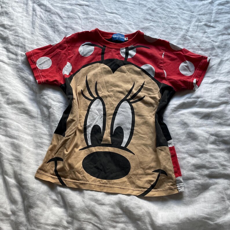 May include: A red t-shirt with white polka dots and a large Minnie Mouse graphic. The shirt features a tan face, black eyes, and a black nose. The sleeves are red with white polka dots. The t-shirt is made of a soft material.