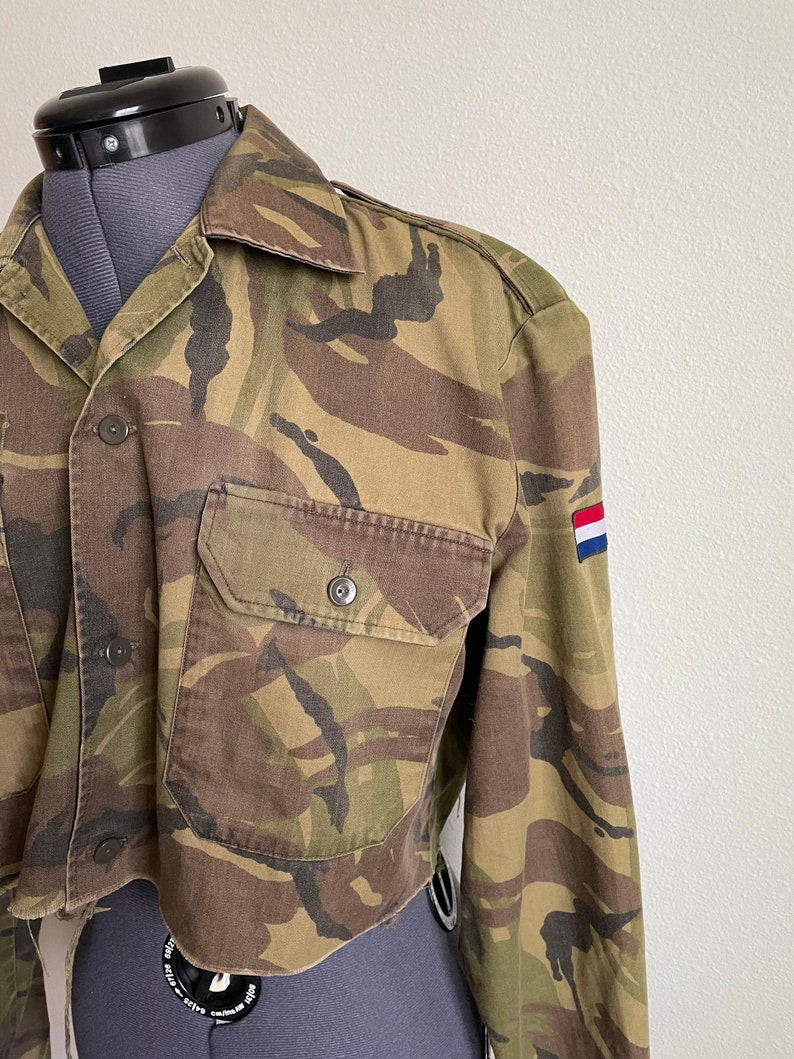 Może przedstawiać: Kr&oacute;tka kurtka w kamuflaż z długimi rękawami i naszywką na rękawie w kolorach flagi holenderskiej.