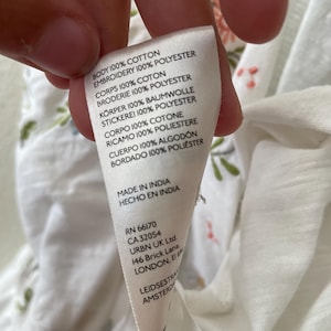 May include: A white fabric tag with text in multiple languages, including English, French, and German. The tag lists the fabric composition as 100% cotton and 100% polyester. The tag also includes the text "MADE IN INDIA" and a company address.