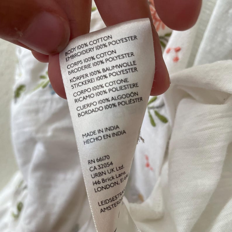 May include: A white fabric tag with text in multiple languages, including English, French, and German. The tag lists the fabric composition as 100% cotton and 100% polyester. The tag also includes the text "MADE IN INDIA" and a company address.
