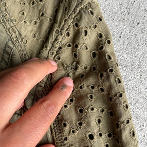 Pode incluir: Close-up de tecido verde-oliva com bordado de ilh&oacute;s intrincado. O tecido &eacute; texturizado e detalhado, com uma costura vis&iacute;vel e uma m&atilde;o tocando o material. O design de ilh&oacute;s cria um visual delicado e arejado.